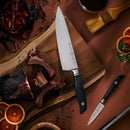 Overhead view of a cutting board with cooked ribs, the Messermeister Blacksmith Paring Knife - 3.5", a bowl of dried chilies, peppercorns, sliced blood oranges, a basting brush, and a cup of dark liquid on a brown surface.
