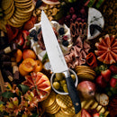 The Messermeister Blacksmith Chef's Knife - 8" rests on a charcuterie board topped with crackers, fruits, olives, meats, cheeses, and flowers—all artfully arranged for a vibrant and abundant presentation.