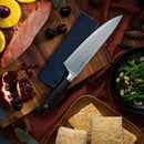 A Messermeister Midnight Navy Leather Magnetic Edge-Guard - 8.5" protects a chef’s knife on a wooden board next to sliced ham with pineapple, cherries, pomegranate, green beans with almonds, biscuits, and decorative pine cones.