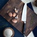 The Messermeister Blacksmith Chef's Knife - 8" rests on a wooden cutting board with three small potatoes, garlic cloves, fresh herbs, and flour. A pepper grinder is visible nearby. Crafted from German stainless steel by Messermeister.