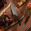 The Messermeister Blacksmith Kullens Santoku Knife - 6.5" rests on a wooden cutting board beside cooked ribs, peppercorns, orange slices, parsley, a paring knife, a pastry brush, and a pot with dried chilies.