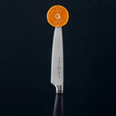 A Messermeister Blacksmith Next Level Block Set - 5 Piece knife stands upright against a dark background, its black handle supporting a round orange slice balanced delicately on the high-carbon stainless steel tip.