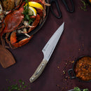 The Messermeister Chef April Bloomfield Carbon Chef's Knife - 6.5" rests on a dark surface beside a plate with cooked crab, lemon wedges, herbs, crab crackers, a bowl of stew, and scattered seasonings.