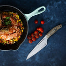 A skillet with shrimp and diced vegetables sits on a blue surface next to cherry tomatoes and a Messermeister Chef April Bloomfield Carbon Chef's Knife - 6.5".