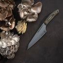 The Messermeister Chef April Bloomfield Carbon Chef's Knife - 6.5" with a wooden handle rests on a dark surface beside oyster mushrooms in white, beige, and brown tones.