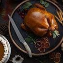 A roasted whole turkey on a round slate tray is garnished with herbs, dried orange slices, cranberries, and wheat stalks. The Messermeister Carbon Scalloped Knife - 9" rests nearby, along with pinecones, autumn leaves, and a pumpkin pie topped with whipped cream.