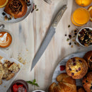 A breakfast spread features pastries, muffins, fruit, a latte, orange juice, and the Messermeister Carbon Next Level Block Set - 5 Piece knife by Messermeister on a light wooden table with crumbs and seeds scattered around.