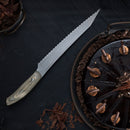 The Messermeister Carbon Scalloped Knife - 9" with a wooden handle rests beside a round chocolate cake topped with swirls of frosting and chocolate shavings on a dark tabletop.