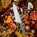 A Messermeister Custom Blue Chef's Knife - 8" rests on a charcuterie board topped with crackers, cheeses, meats, olives, apricots, strawberries, assorted fruits, and is beautifully surrounded by decorative flowers.