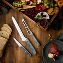 The Messermeister Custom Blue Scalloped Offset Knife - 8" rests on a wooden table with bread slices, beside two other blue-handled knives. In the top right, a large wooden tray displays assorted cheeses, fruits, crackers, and charcuterie.