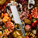 The Messermeister Custom Blue Nakiri Knife - 6.5" rests on a platter topped with vibrant fruits, cheeses, crackers, charcuterie, nuts, olives, and flowers for an abundant display.