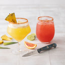 Two cocktails—a yellow one with pineapple and chili rim, a red one on ice—sit on a light surface next to the Messermeister Custom Blue Paring Knife - 3.5", lime slices, pineapple pieces, and an orange wedge.
