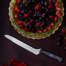 A fruit tart topped with assorted berries and cherries sits beside a Messermeister Custom Scalloped Offset Knife - 8" on a dark surface, with extra cherries and rose petals scattered nearby.