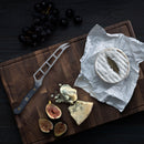 A dark surface displays a wooden board with soft round cheese in paper, blue cheese wedges, figs, dark grapes, and the Messermeister Custom Cheese & Tomato Knife - 5" by Messermeister.
