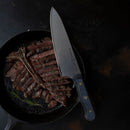 A sliced cooked steak in a cast iron skillet with a sprig of rosemary, accompanied by a large chef’s knife from the Messermeister Custom Next Level 5-Piece Magnetic Knife Block Set resting across the skillet on a dark background.