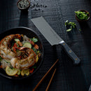 A plate of grilled shrimp on noodles with vegetables sits beside the Messermeister Custom Nakiri Knife - 6.5", a bowl of sliced green onions, a bowl of sesame seeds, and wooden chopsticks on a dark textured table.