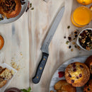 A Messermeister Custom Next Level 5-Piece Magnetic Knife Block Set from Messermeister’s precision cutting collection rests on a light wooden table, surrounded by pastries, orange juice, a fork, and bowls filled with mixed nuts and seeds. Crumbs and fruit are scattered among the breakfast spread, creating an inviting scene that highlights both the organized kitchen storage and premium cutlery.