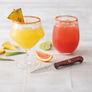 Two cocktails sit on a tiled surface: one yellow with a pineapple slice and chili rim, the other pink with a chili rim. Nearby, the Messermeister Custom Red Paring Knife - 3.5" rests beside sliced pineapple, lime, and orange.