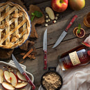 A rustic table displays an apple pie, sliced apples, cinnamon sticks, brown sugar, a whiskey bottle and glass, plus three razor-sharp knives from the Messermeister Custom Red 4 Piece Steak Knife Set arranged among the ingredients.