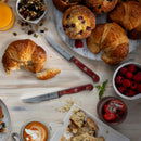 A breakfast scene with croissants, muffins, a bowl of raspberries, cake, coffee, orange juice, and razor-sharp Messermeister Custom Red 4 Piece Steak Knife Set on a light table.