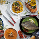 A top view of a table set with seafood dishes—whole grilled fish, shrimp with salsa, soup, gnocchi, oysters, crab claw, cherry tomatoes—and three sharp Messermeister Custom Red 4 Piece Steak Knife Set knives.