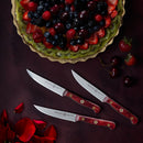 A berry tart with strawberries, blueberries, raspberries, and cherries sits on a green plate. Below are three Messermeister Custom Red steak knives and scattered cherries, strawberries, and rose petals on a dark surface.