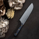 The Messermeister Custom Kullens Santoku Knife - 7" with a dark handle rests on a dark surface beside clusters of oyster mushrooms in beige, cream, and light brown.