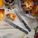 The Messermeister Custom Steak Knife Set – 4 Piece, featuring razor-sharp steel blades, rests on a light wooden surface with croissants, a cranberry muffin, mixed nuts and a marble tray for an inviting brunch display.