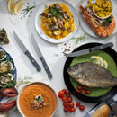 A selection of seafood—whole fish in a skillet, shrimp, gnocchi, lobster, oysters, tomato soup, bread, and cherry tomatoes—is beautifully arranged on a white table with the Messermeister Custom Steak Knife Set - 4 Piece by Messermeister.