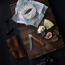 A cheese board with assorted cheeses, figs, and grapes is paired with a Messermeister Folding Steak Knife from the 4 Piece Set in a Leather Roll by Messermeister, displayed on a dark wooden surface.