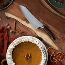 A pumpkin pie with whipped cream sits on a wooden board next to the Messermeister Kawashima Magnet Block Set - 5 Piece, surrounded by dried orange slices, cinnamon sticks, pecans, and a rust-colored cloth.
