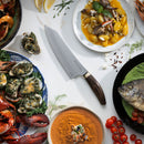 A variety of gourmet dishes surround the Messermeister Kawashima Magnet Block Set - 5 Piece by Messermeister, beautifully arranged on a white table with herbs, lemon wedges, and cherry tomatoes for garnish.