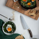 A bowl and a skillet with sautéed greens and sunny-side-up eggs, garnished with seeds and herbs, sit beside pita bread, a Messermeister Kawashima Chef's Knife - 8", a fork, and a green napkin on a white table.