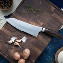 The Messermeister Kawashima Chef's Knife - 8" by Messermeister, with an ergonomic handle, sits on a wooden cutting board surrounded by garlic cloves, small potatoes, peppercorns, thyme, and flour on a blue surface.