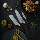 Three kitchen knives, featuring the Messermeister Kawashima Nakiri Knife - 6.5", rest on a dark surface beside asparagus, tomatoes, and herbs. A skillet with sautéed carrots, onions, and potatoes completes the inspiring culinary scene.