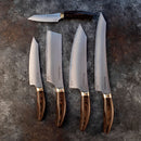 Five Messermeister kitchen knives, including the Kawashima Nakiri Knife - 6.5", with wooden handles, are arranged on a dark stone surface in order of size and shape, from the smallest at the top to largest at the bottom right.