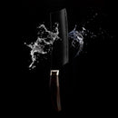 The Messermeister Kawashima Nakiri Knife - 6.5" by Messermeister features a dark handle and a sharp SG2 powdered steel blade, shown against a black background with water splashes scattering dramatically around it.