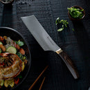 A 6.5" Messermeister Kawashima Nakiri knife with a wooden handle lies on a dark textured surface beside a bowl of vegetables and shrimp, sliced green onions in a small bowl, and wooden chopsticks.