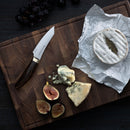 A wooden cutting board holds a Messermeister Kawashima Paring Knife - 3.5", brie on parchment, blue cheese wedges, figs, and black grapes, all attractively arranged on a dark surface.