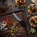 A Messermeister Kawashima Paring Knife - 3.5" by Messermeister rests on a cutting board with diced veggies, sliced onions and peppers, and cooked meat, while tacos topped with meat, veggies, limes, and spices sit nearby on a dark surface.