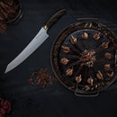 A glossy chocolate cake topped with frosting and chocolate shavings sits on a tray. Next to it lies the Messermeister Kawashima Slicer - 10", crafted with SG2 powdered steel and renowned Japanese craftsmanship, on a dark wooden surface.