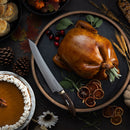 A roasted whole turkey on a round tray with herbs, cranberries, and dried orange slices, surrounded by pumpkin pie, biscuits, pinecones, corn, and autumn leaves. The Messermeister Kawashima Slicer - 10" rests beside the turkey.