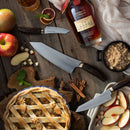 A rustic table with apples, an apple pie, sliced apples, the Messermeister Kawashima 8" Chef’s Knife, two additional kitchen knives, a bottle of Makers Mark whiskey, brown sugar in a dish, cinnamon sticks, and dried citrus slices.