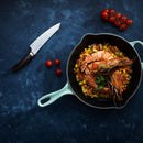 A skillet of shrimp and diced veggies rests on a blue surface beside the Messermeister Kawashima Utility Knife - 6", loose cherry tomatoes, and a vine of cherry tomatoes—ideal for kitchen prep.