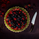 A vibrant fruit tart sits beside the Messermeister Kawashima Magnet Block Set - 5 Piece by Messermeister, with a SG2 steel knife on a dark surface scattered with berries, rose petals, and chocolate shavings.