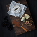 A wooden board displays a round wheel of soft cheese on parchment, a wedge of blue cheese, sliced figs, dark grapes, and a cheese knife with the Messermeister Saddle Brown Leather Magnetic Edge-Guard - 4".