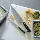 Two kitchen knives, including the Messermeister Meridian Elite Spear Point Paring Knife - 4", rest on a white cutting board among chopped onions, carrots, zucchini, and tomatoes. A whole tomato and containers of diced veggies sit nearby.
