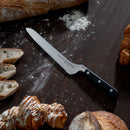 A Messermeister Meridian Elite Magnet Block Set - 8 Piece bread knife with a black handle rests on a dark wooden table among breads, pastries, and scattered flour, showcasing this premium German steel kitchen knife collection.
