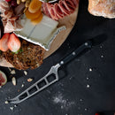 A charcuterie board with meats, cheese, dried apricots, strawberries, multigrain bread, and the Messermeister Meridian Elite Cheese & Tomato Knife - 5", rests on a dark surface surrounded by bread and scattered crumbs.