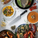 A top-down view of a table featuring seafood dishes, lemon slices, cherry tomatoes, and a Messermeister Meridian Elite Chef's Knife - 8" at the center.